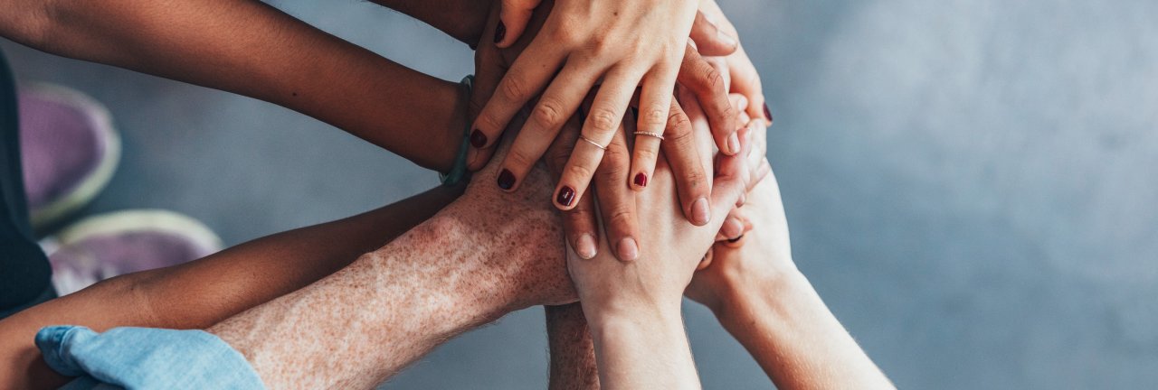 Zu sehen ist eine Gruppe von Menschen, die ihre Hände in der Mitte aufeinander. Es handelt sich um eine Draufsicht, die die Hände unter Unterarme zeigt. 