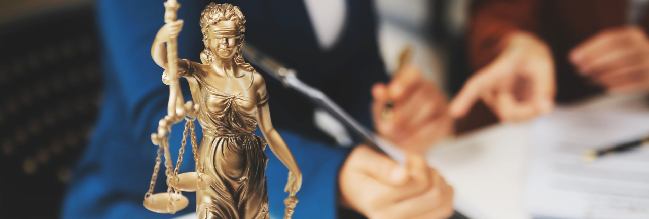 Eine Justitia-Figur steht auf dem Tisch; dahinter arbeiten zwei Personen an Dokumenten.