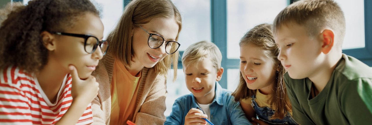 Vier Grundschulkinder sitzen zusammen mit einer Lehrerin am Tisch. Sie zeigt ihnen etwas auf den Papieren, die vor ihnen liegen.