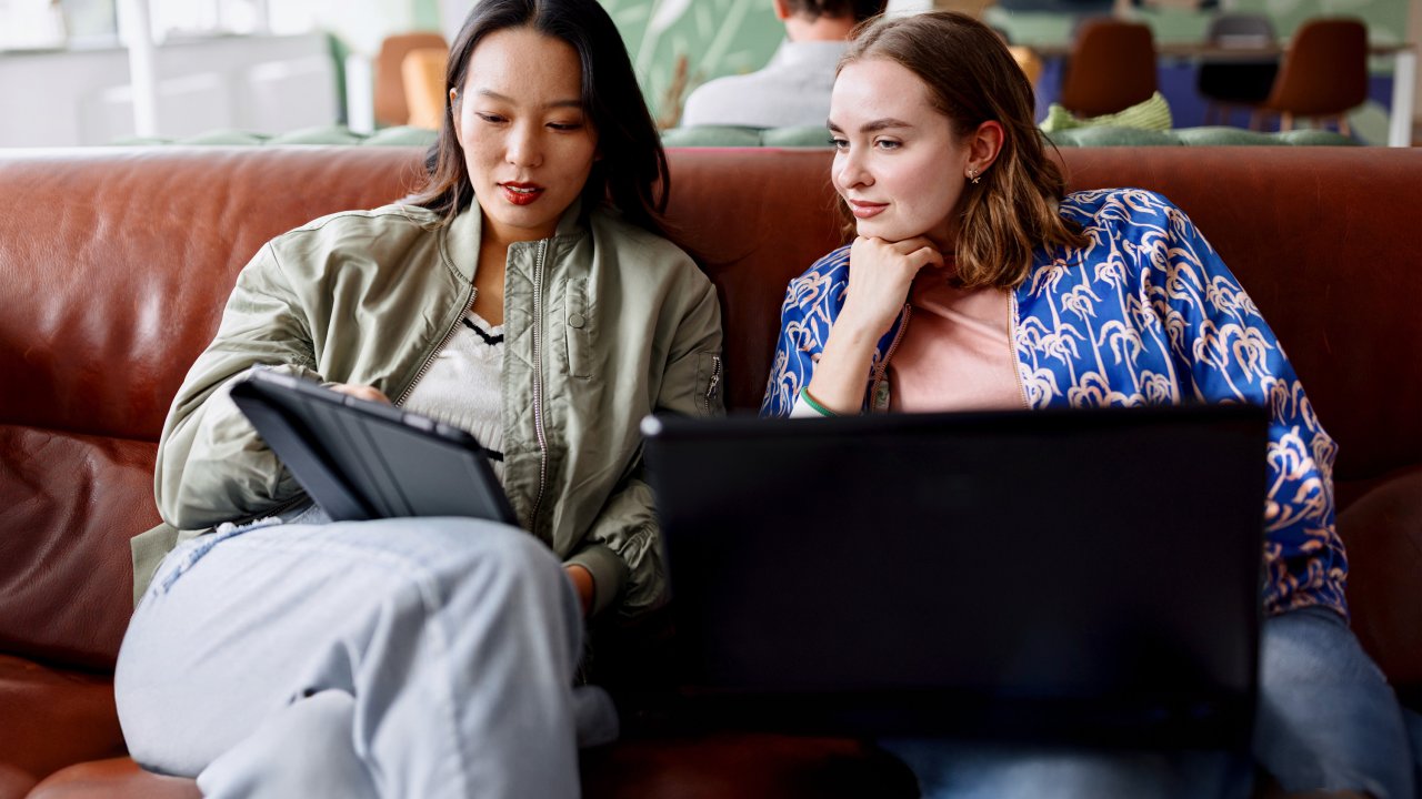 Zwei Frauen sitzen auf einem Sofa; die eine Frau hat einen Laptop auf dem Schoß, die andere hat ein Tablet in der Hand.