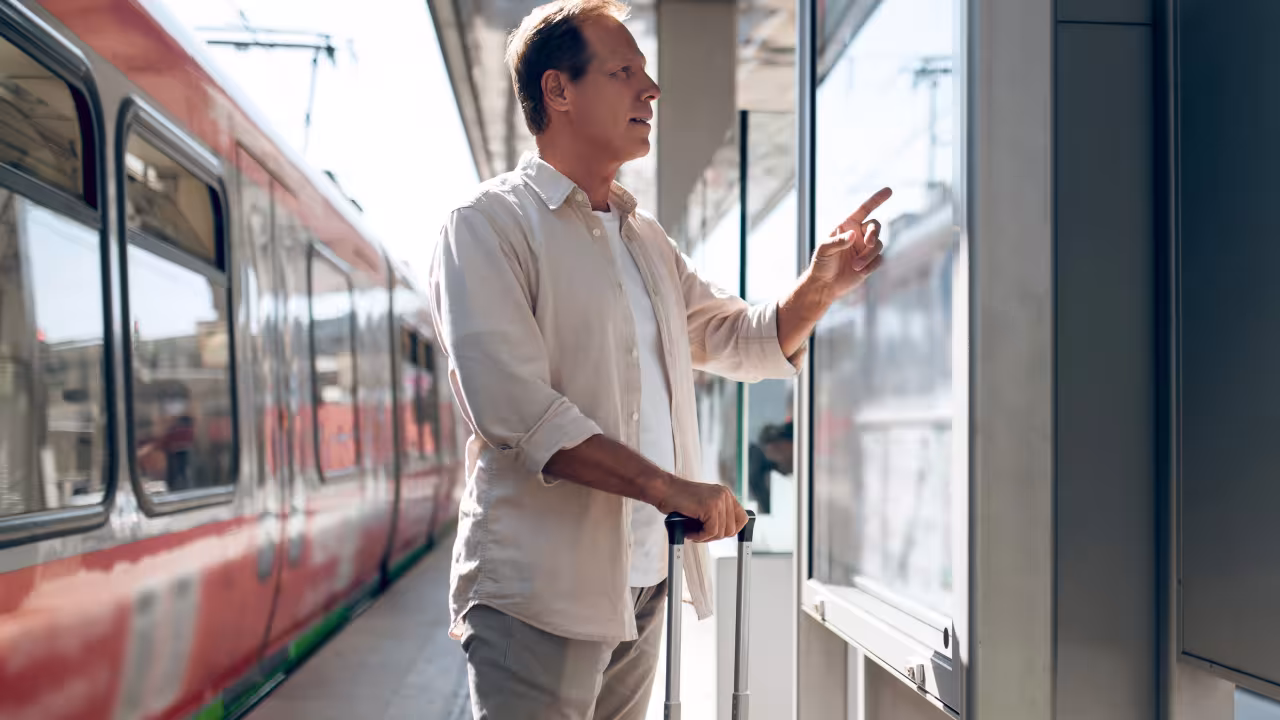 Mann steht an einem Bahnsteig und guckt auf einen Aushang; hinter ihm ist ein Zug zu sehen