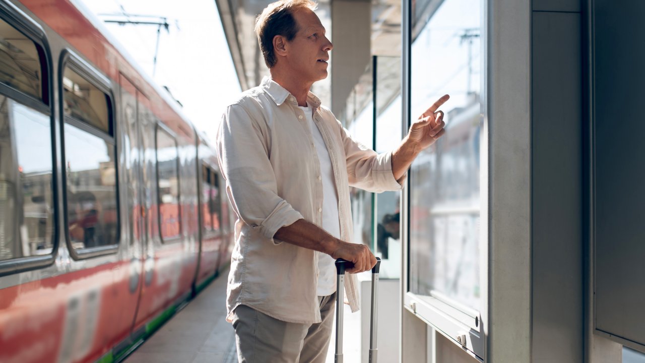 Mann steht an einem Bahnsteig und guckt auf einen Aushang; hinter ihm ist ein Zug zu sehen