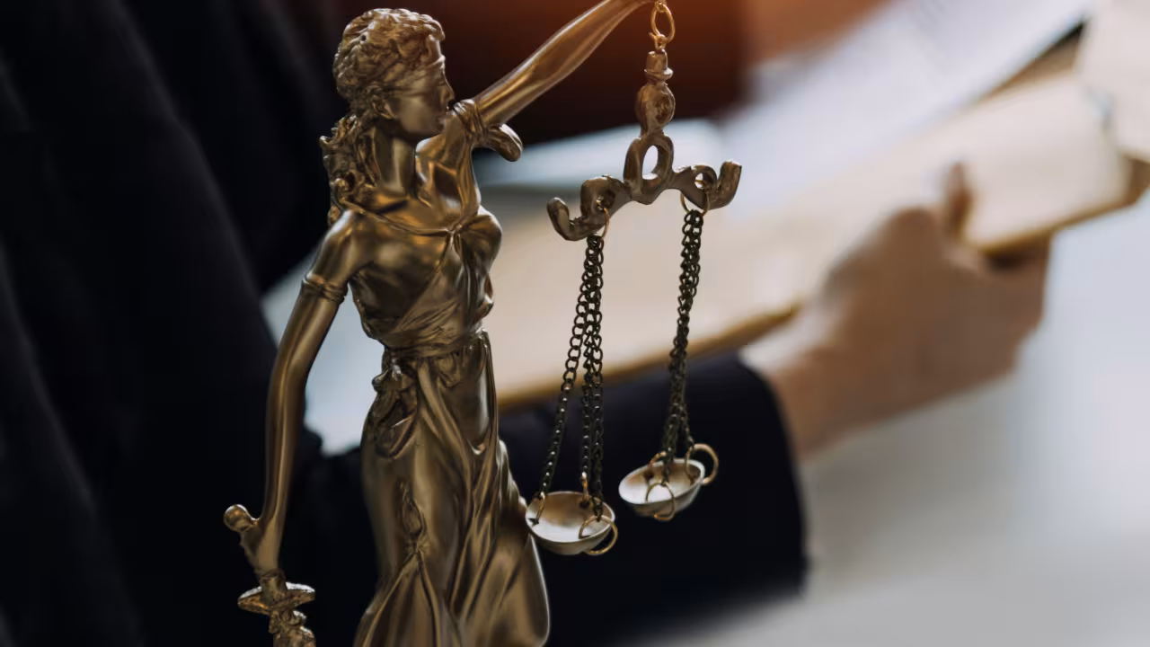 Goldene Justitia-Figur steht auf einem Tisch; dahinter ist eine Person zu erkennen, die Dokumente in der Hand hält