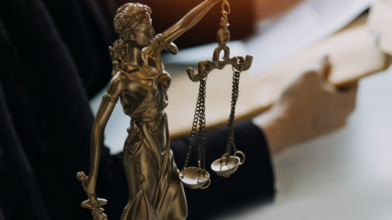 Goldene Justitia-Figur steht auf einem Tisch; dahinter ist eine Person zu erkennen, die Dokumente in der Hand hält