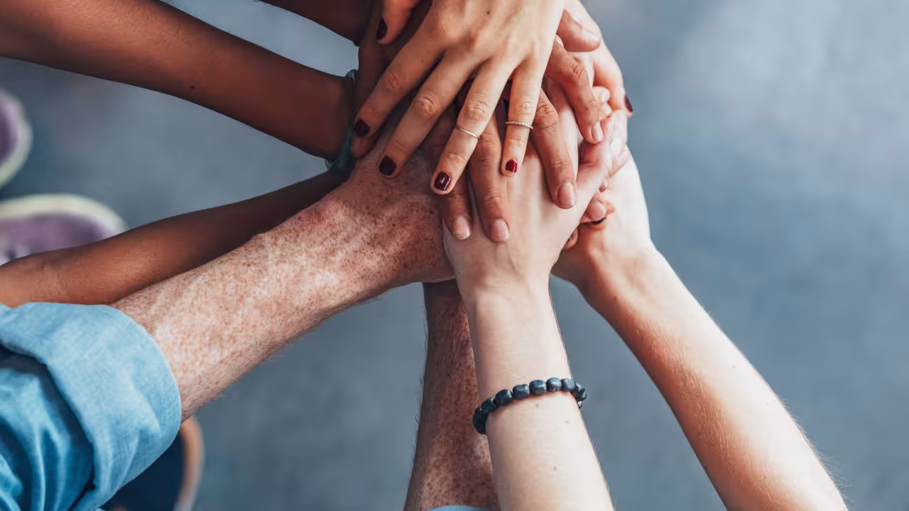 Zu sehen ist eine Gruppe von Menschen, die ihre Hände in der Mitte aufeinander. Es handelt sich um eine Draufsicht, die die Hände unter Unterarme zeigt. 