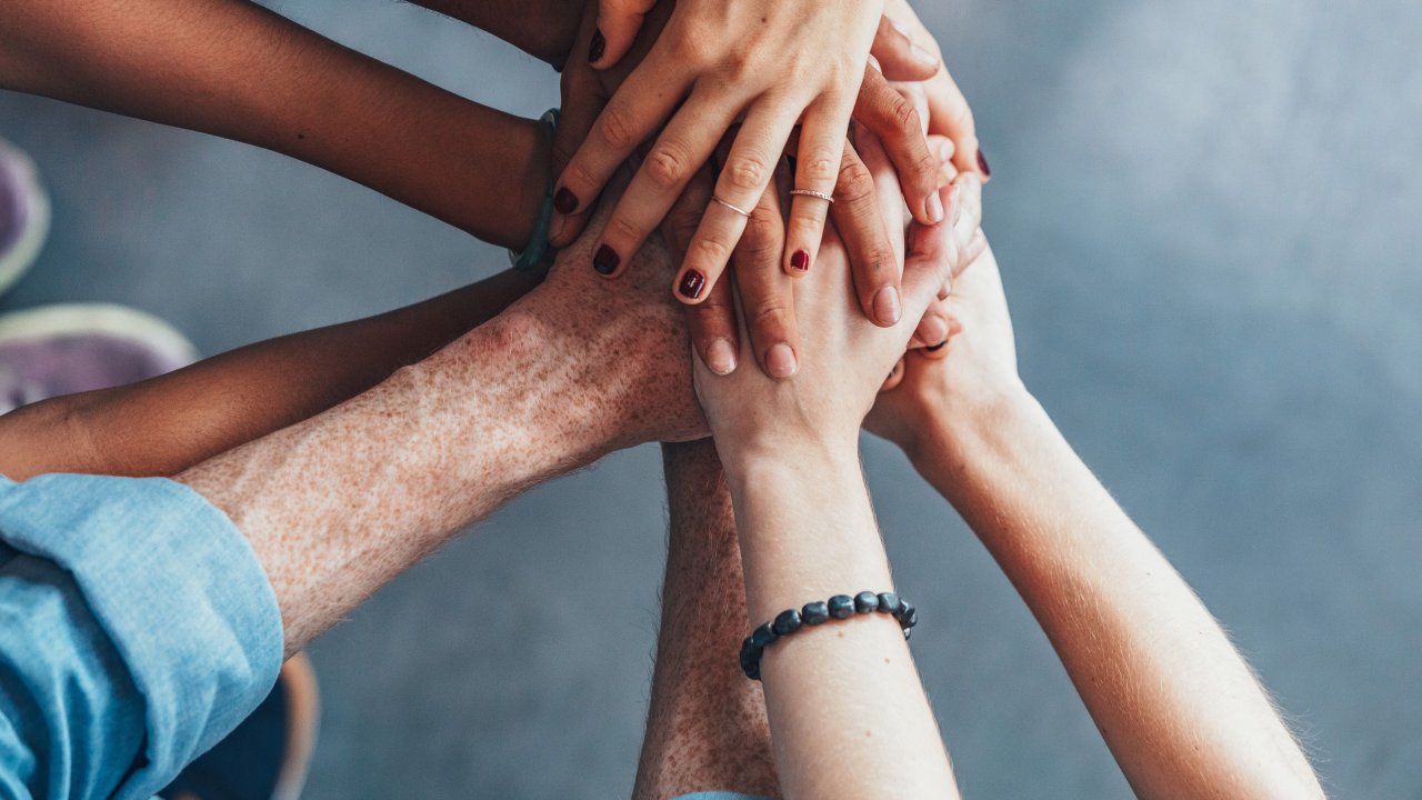 Zu sehen ist eine Gruppe von Menschen, die ihre Hände in der Mitte aufeinander. Es handelt sich um eine Draufsicht, die die Hände unter Unterarme zeigt. 