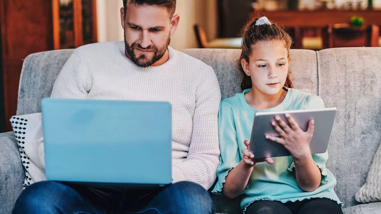 Ein Mann und ein Kind sitzen nebeneinander auf dem Sofa. Der Mann hat einen Laptop auf dem Schoß, das Mädchen ein Tablet in der Hand.