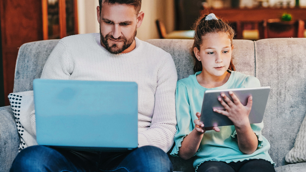 Ein Mann und ein Kind sitzen nebeneinander auf dem Sofa. Der Mann hat einen Laptop auf dem Schoß, das Mädchen ein Tablet in der Hand.