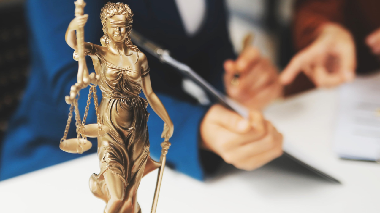 Eine Justitia-Figur steht auf dem Tisch; dahinter arbeiten zwei Personen an Dokumenten.