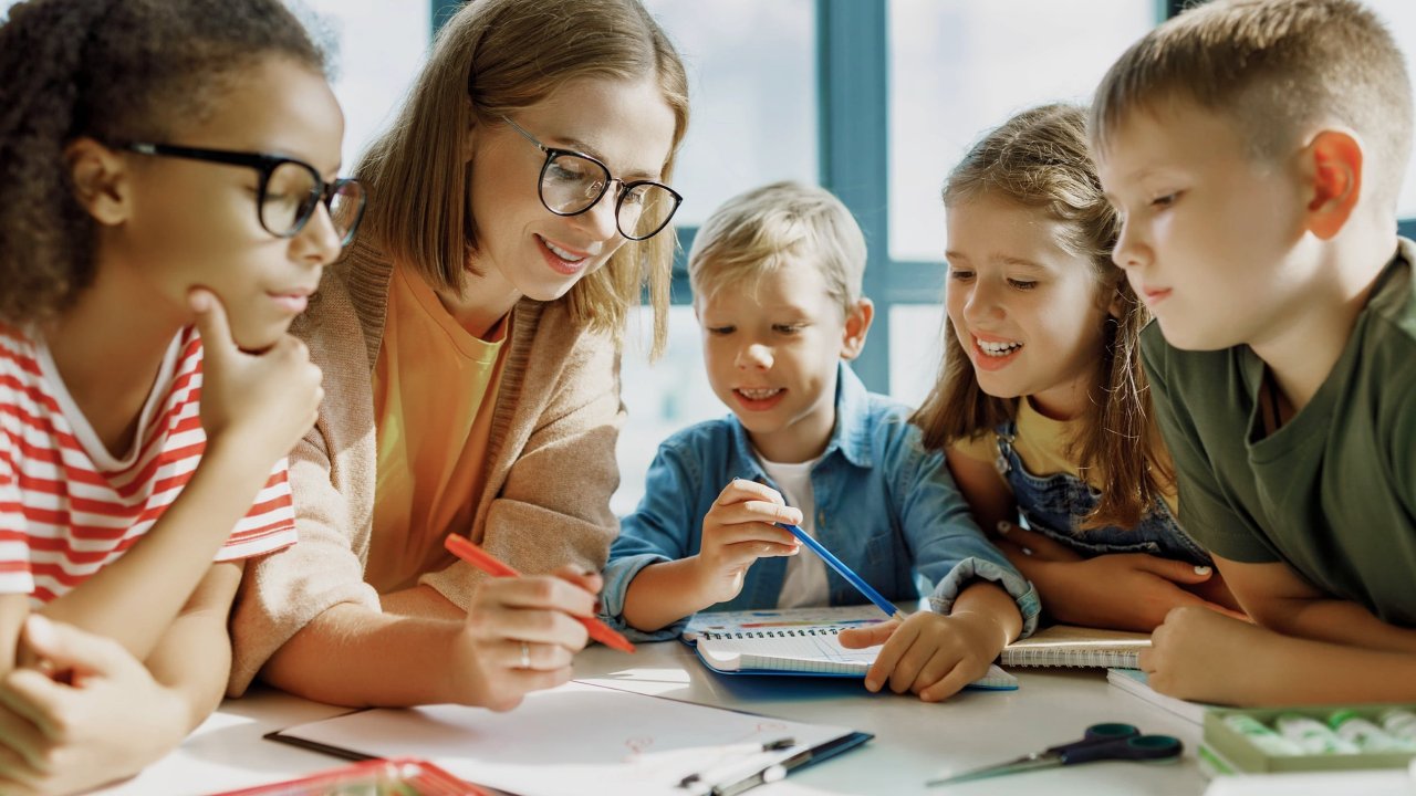 Vier Grundschulkinder sitzen zusammen mit einer Lehrerin am Tisch. Sie zeigt ihnen etwas auf den Papieren, die vor ihnen liegen.