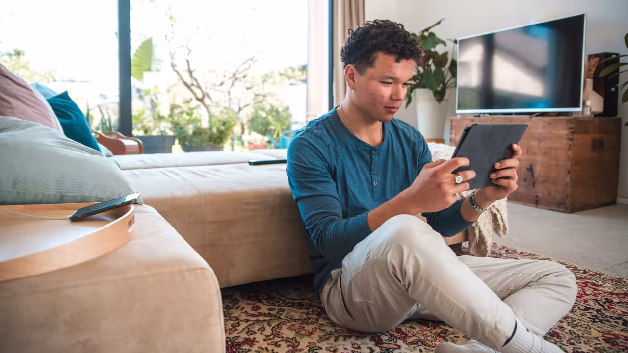 Ein Jugendlicher sitzt auf dem Teppich vor dem Sofa mit einem Tablet in der Hand.