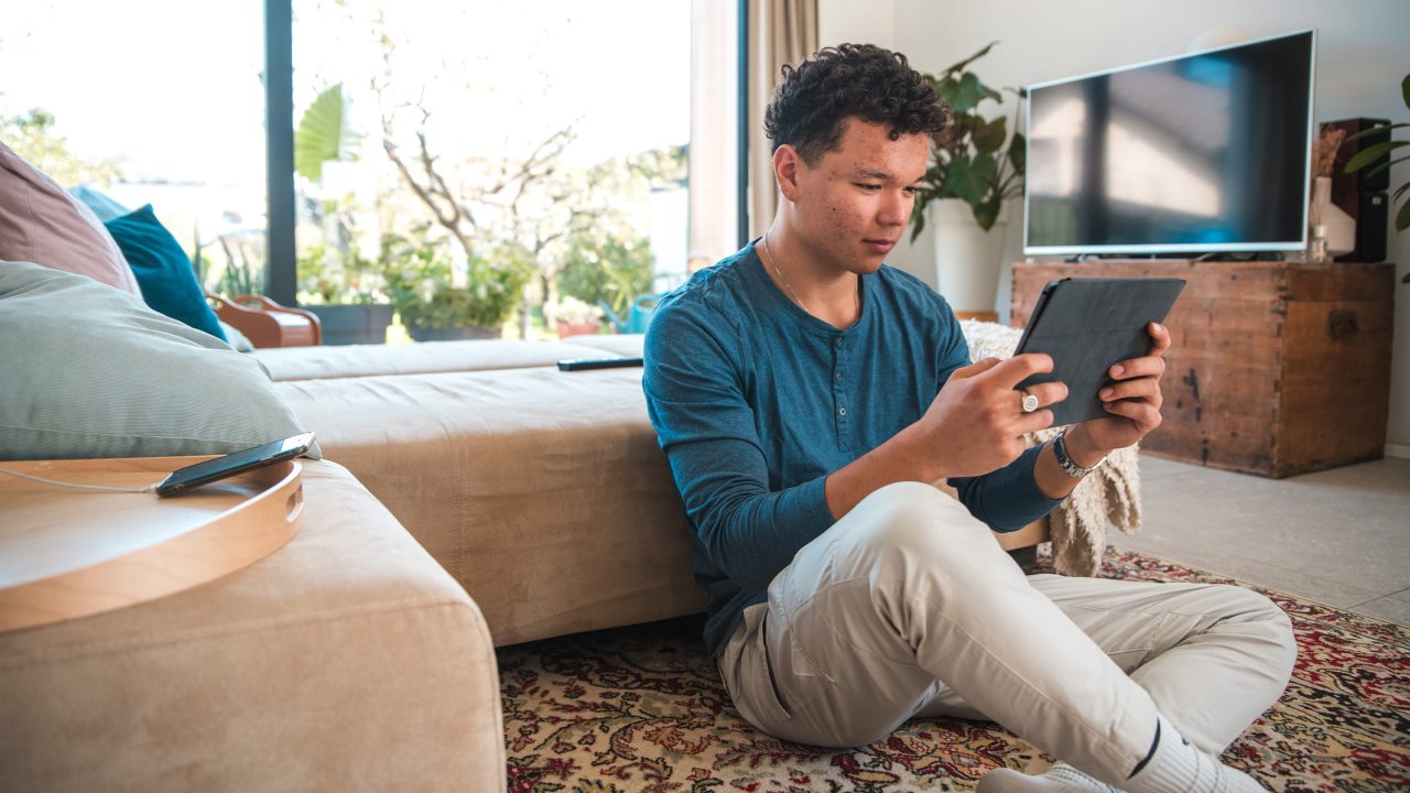Ein Jugendlicher sitzt auf dem Teppich vor dem Sofa mit einem Tablet in der Hand.