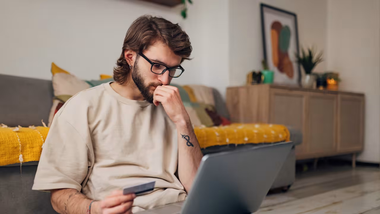 Ein junger Mann sitzt vor dem Sofa auf dem Boden mit dem Laptop auf dem Schoß und einer Geldkarte in der Hand.