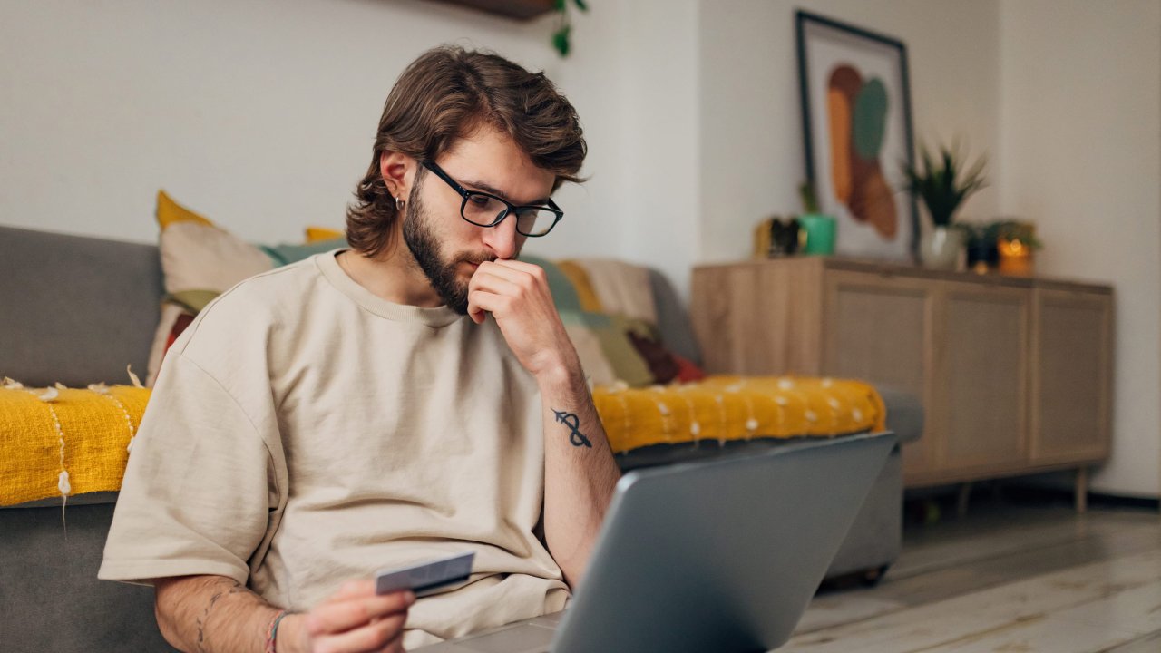 Ein junger Mann sitzt vor dem Sofa auf dem Boden mit dem Laptop auf dem Schoß und einer Geldkarte in der Hand.