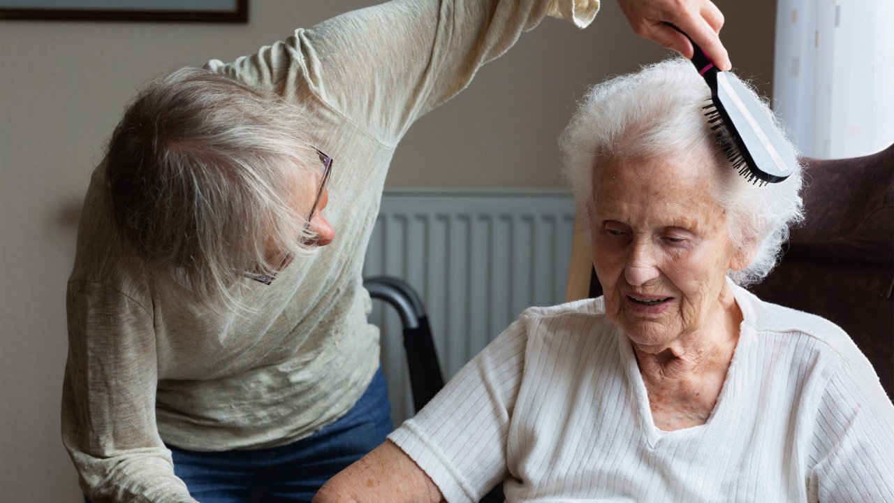 Eine Frau kämmt einer Seniorin die Haare, die in einem Sessel sitzt.