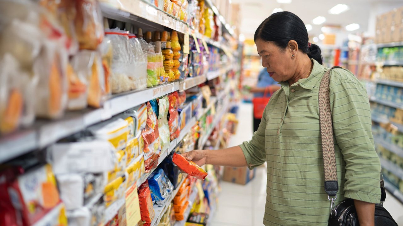 Eine Frau steht im Supermarkt vorm Regal und schaut prüfend auf ein Produkt, das sie in der Hand hält.