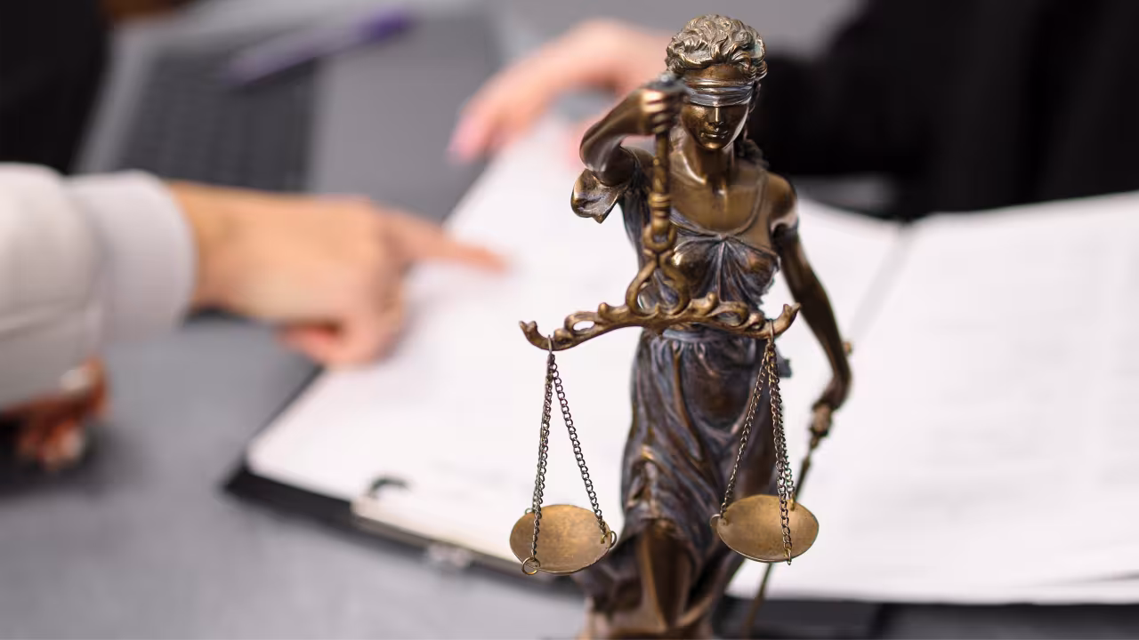Eine Justitia-Figur steht auf dem Schreibtisch; hinter hier sind zwei Hände über Dokumenten zu sehen.