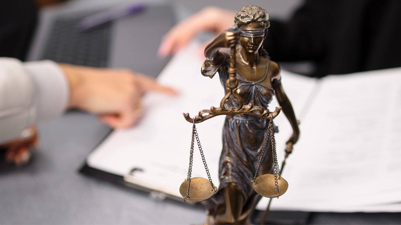 Eine Justitia-Figur steht auf dem Schreibtisch; hinter hier sind zwei Hände über Dokumenten zu sehen.