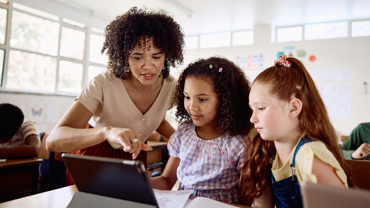 Eine Lehrerin erklärt zwei Kindern etwas an einem Tablet.