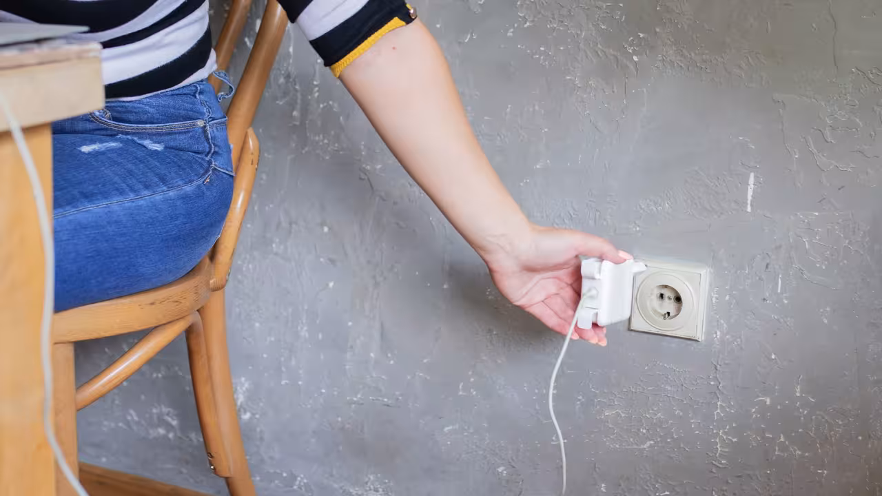Eine Person sitzt am Tisch, auf dem ein Laptop steht, und steckt hinter sich ein Ladegerät in die Steckdose.