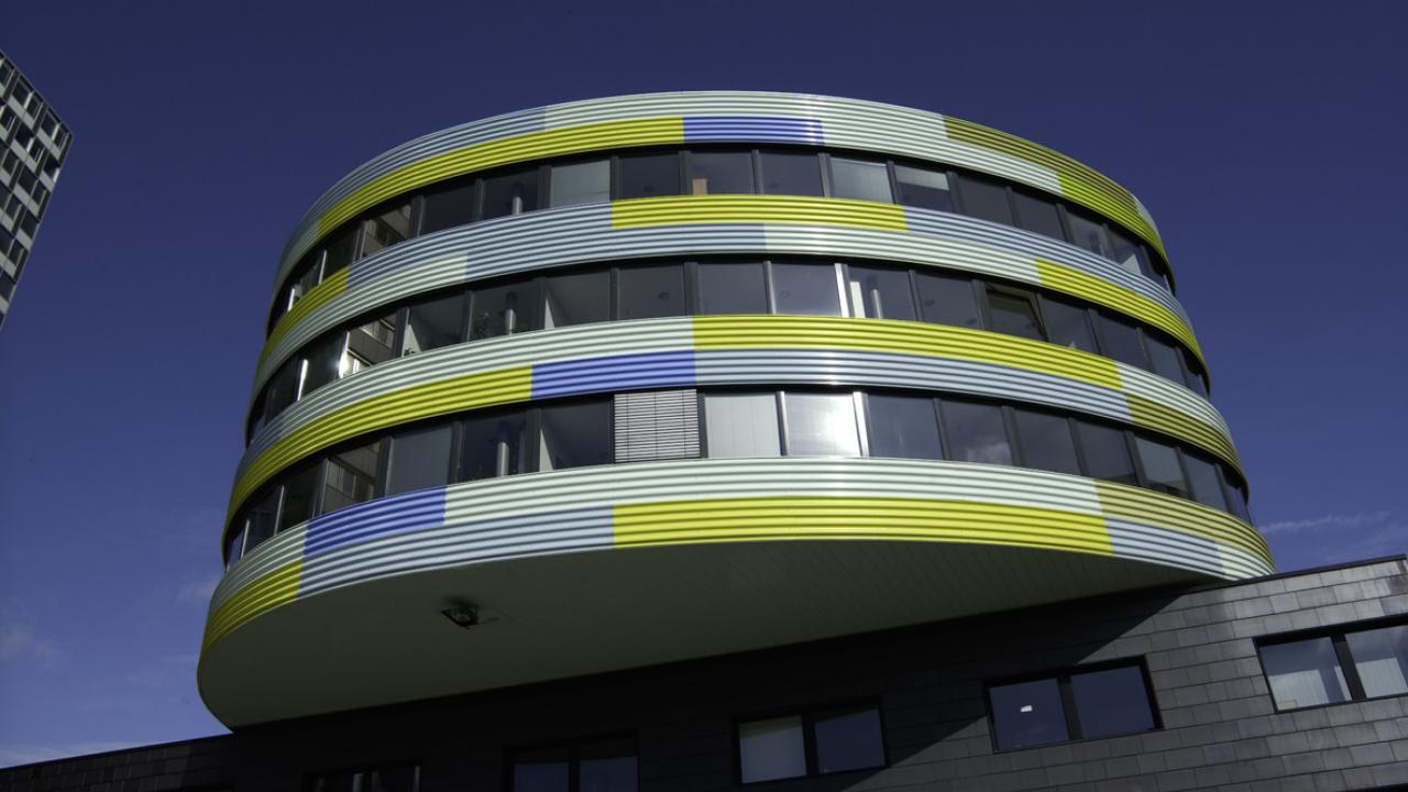 Zu sehen ist ein rundes dreistöckiges Gebäudes mit umlaufenden Fenstern. Die Fassadengestaltung ist neongelb und hebt sich um blauen Himmel ab. Bei der sogenannte Pillbox handelt es sich um ein Gebäudeteil, in dem der vzbv seine Büroräume hat. 